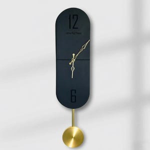 Op de afbeelding: Een moderne, zwarte wandklok met goudkleurige wijzers en een slinger. De klok heeft de cijfers 12 en 6, met de tekst "Urris by Nooz". De slinger heeft een rond, goudkleurig gewicht.
