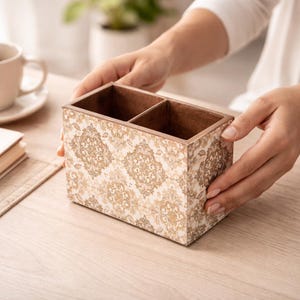 May include: A decorative wooden desk organizer with two compartments. The exterior features a cream-colored background with a gold damask pattern. The interior and top edges are a dark brown color. The organizer is held by a person's hands.