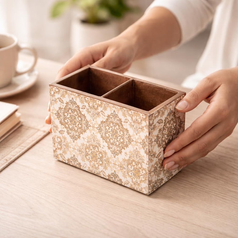 May include: A decorative wooden desk organizer with two compartments. The exterior features a cream-colored background with a gold damask pattern. The interior and top edges are a dark brown color. The organizer is held by a person's hands.