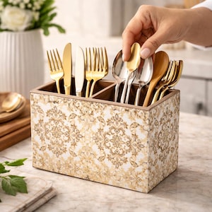 May include: A wooden utensil holder with a white and gold damask pattern. The holder is divided into three sections and contains various silverware, including gold forks, knives, and spoons. A hand is placing a gold spoon into the holder.