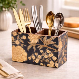 May include: A wooden utensil holder with a black background and gold floral design. The holder is divided into four compartments, each holding different silverware: gold knives and forks, and silver and gold spoons.