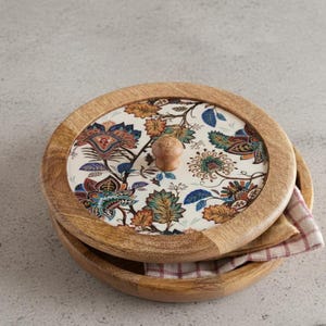 May include: A wooden serving bowl with a lid, featuring a floral pattern in blue, orange, and green on a white background. The lid has a small wooden knob. A checkered napkin is inside the bowl. The bowl is made of wood.