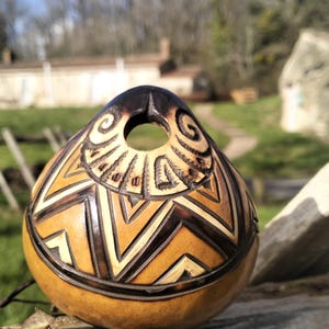 May include: A handcrafted gourd with a dark brown top and intricate geometric patterns carved into its tan surface. The gourd features a circular opening and a decorative border, showcasing a natural, earthy aesthetic.