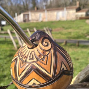 May include: A handcrafted gourd with intricate geometric carvings and a stainless steel straw. The gourd features a natural brown color with lighter wood accents, and a dark brown top. The background shows a sunny outdoor scene.