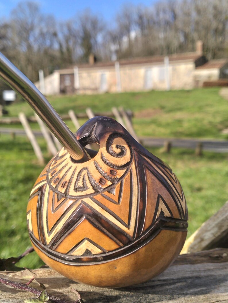 May include: A handcrafted gourd with intricate geometric carvings and a stainless steel straw. The gourd features a natural brown color with lighter wood accents, and a dark brown top. The background shows a sunny outdoor scene.