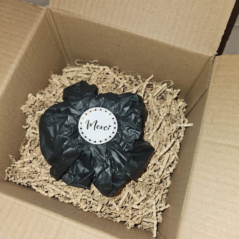 May include: A cardboard box filled with brown paper shreds. Inside, black tissue paper is wrapped around an item, with a white sticker that says "Merci" in cursive, surrounded by small heart shapes.