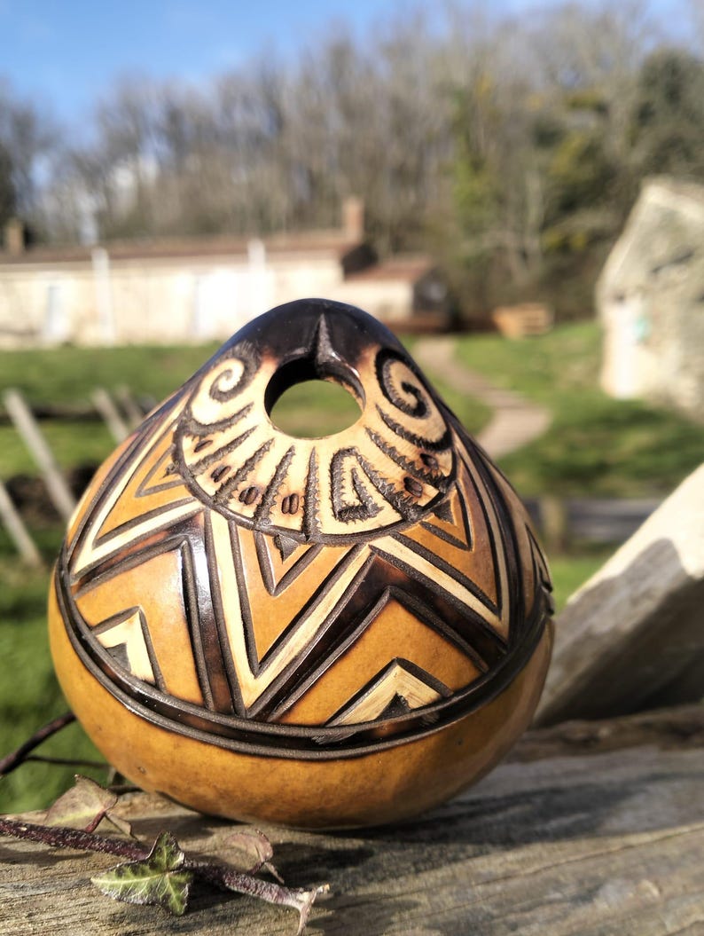 May include: A handcrafted gourd with a dark brown top and intricate geometric patterns carved into its tan surface. The gourd features a circular opening and a decorative border, showcasing a natural, earthy aesthetic.