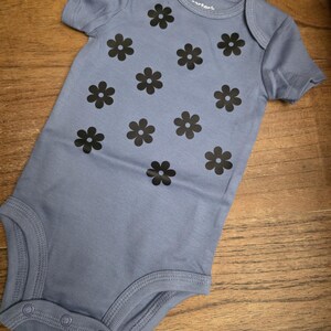 Puede incluir: Un body de beb&eacute; azul claro con mangas cortas y cierre de botones a presi&oacute;n. El body est&aacute; decorado con motivos de flores negras. La prenda est&aacute; hecha de una tela suave.