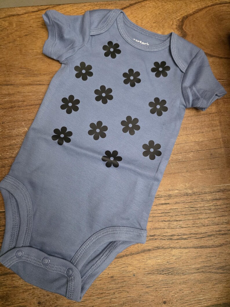 Puede incluir: Un body de beb&eacute; azul claro con mangas cortas y cierre de botones a presi&oacute;n. El body est&aacute; decorado con motivos de flores negras. La prenda est&aacute; hecha de una tela suave.