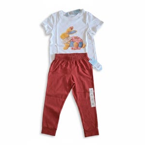 Op de afbeelding: Een set bestaande uit een wit T-shirt en een roestkleurige broek. Het T-shirt heeft een patchwork konijnontwerp met gele, blauwe en roze accenten. De broek heeft een elastische tailleband en een aangehecht label.
