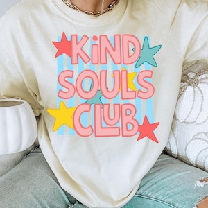 Może przedstawiać: Kremowa bluza z napisem "KIND SOULS CLUB" w kolorze różowym, obramowanym na niebiesko i ozdobionym kolorowymi gwiazdami. Tekst umieszczono na jasnoniebieskim tle w paski.