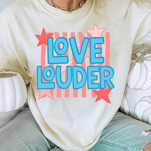 Może przedstawiać: Kremowa koszulka z napisem "LOVE LOUDER" w kolorze niebieskim, obramowanym na biało i ozdobionym czerwonymi gwiazdami. Tekst umieszczono na tle różowych, pionowych pasków. Swobodny, wygodny styl.