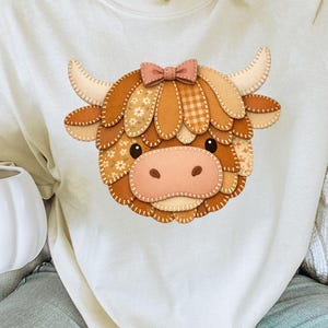 May include: Cream-coloured sweatshirt with a detailed appliqué of a Highland cow's face. The cow has a patchwork design in shades of brown, tan, and peach, with a pink bow. The sweatshirt is a casual, comfortable style.