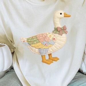 Puede incluir: Sudadera color crema con un aplique de ganso. El ganso tiene un diseño de patchwork con estampados florales y de cuadros en colores pastel. Tiene un cuello con volantes verdes y un lazo a cuadros. El pico y las patas del ganso son amarillos.