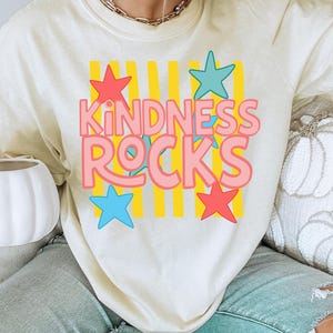 Może przedstawiać: Kremowa bluza z napisem "KINDNESS ROCKS" w kolorze różowym i niebieskim, z czerwonymi i niebieskimi gwiazdami oraz żółtymi paskami. Koszulka jest w stylu casual i wygodnym.