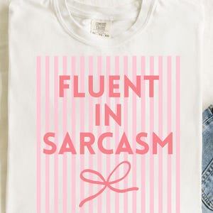 May include: White t-shirt with the phrase "FLUENT IN SARCASM" in pink block letters, set against a background of vertical pink stripes. A pink bow graphic is at the bottom. The shirt is made of a soft material.
