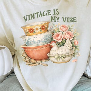 以下が含まれることがあります： 「VINTAGE IS MY VIBE」の文字が緑色でプリントされたクリーム色のスウェットシャツ。積み重ねられたヴィンテージボウル、バスケットに入った鶏、ピンクのバラが描かれています。一番上のボウルにはオレンジと白の花があります。