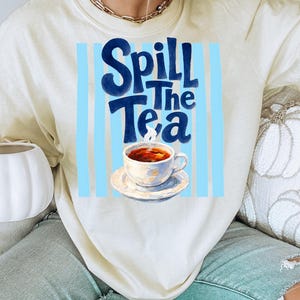 May include: Cream-colored sweatshirt with the phrase "Spill The Tea" in dark blue script. The graphic includes a teacup with tea and steam, set against a light blue striped background. The sweatshirt is a casual, comfortable style.