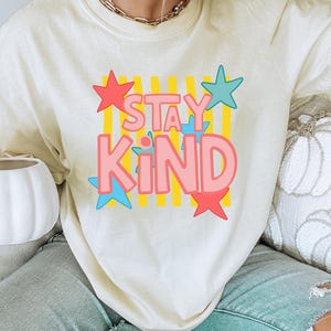 Może przedstawiać: Kremowa koszulka z napisem "STAY KIND" w kolorze różowym, obramowanym na niebiesko, na żółtym tle w paski. Czerwone i niebieskie gwiazdy otaczają tekst. Koszulka jest noszona z niebieskimi dżinsami.
