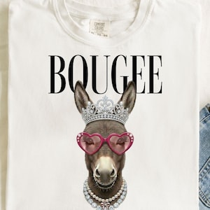 Può includere: T-shirt bianca con la scritta "BOUGEE" a grandi lettere nere. La maglietta presenta una grafica di un asino con corona, occhiali da sole a forma di cuore e collana di perle. L'asino è marrone e grigio.