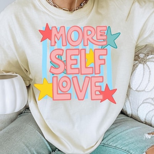 Może przedstawiać: Kremowa koszulka z napisem "MORE SELF LOVE" w kolorze różowym, obramowanym na czerwono, na jasnoniebieskim tle. Projekt zawiera czerwone, żółte i turkusowe gwiazdki. Koszulka to odzież codzienna.