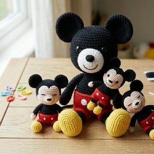 May include: A collection of crocheted Mickey Mouse dolls in different sizes. The dolls are black, red, yellow, and white, with large black ears and smiling faces. They are arranged on a wooden surface near a window.