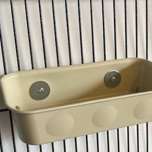May include: A cream-coloured rectangular pet feeder or storage container with three rounded details on the front. The container is attached to a black wire grid with two silver magnetic fasteners.