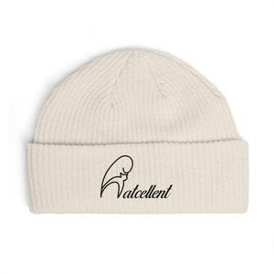 Puede incluir: Gorro de color crema con borde doblado. El gorro presenta un gráfico negro y la palabra "Watcellent" en una fuente estilizada. El gorro está hecho de un material de punto acanalado.