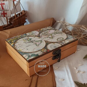 Può includere: Una scatola rettangolare in legno con un design a mappa vintage sul coperchio. La scatola ha una finitura marrone chiaro e una chiusura in metallo. Le parole "Hand Made" e "MartDesigner" sono stampate su un adesivo sul lato. Un modello di nave è sullo sfondo.