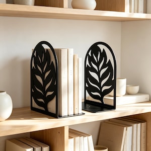 Op de afbeelding: Een paar zwarte metalen boekensteunen met een uitgesneden bladerpatroon en een boogvormige bovenkant. De boekensteunen staan op een houten plank en houden een kleine collectie boeken met lichtgekleurde ruggen vast voor een geordende uitstraling.
