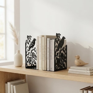 Puede incluir: Un par de sujetalibros de metal negro con un diseño cortado con láser de pájaros posados en ramas de rosas. Los sujetalibros sostienen una fila de libros con los lomos titulados Northern Lights, Heart of Heather y Rustic Paths.