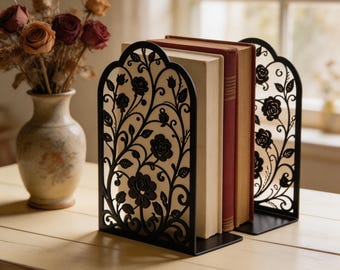 Puede incluir: Un par de sujetalibros de metal negro con un intrincado diseño cortado con láser de rosas trepadoras, hojas y enredaderas. Los sujetalibros arqueados sostienen dos libros de tapa dura con lomos de color crema y rojo sobre una superficie.