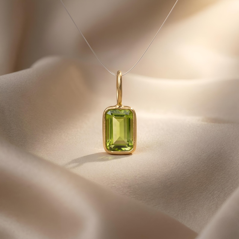 May include: A gold pendant necklace with a rectangular, light green gemstone. The gemstone is set in a gold bezel and suspended from a fine chain. The pendant is shown against a soft, neutral backdrop.
