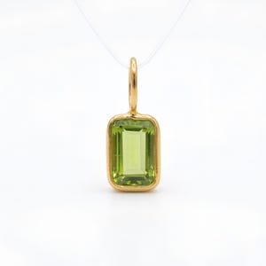 May include: A rectangular peridot pendant with a gold-coloured bezel and bail. The light green peridot stone is set in a gold frame. The pendant hangs from a thin, clear cord, and is set against a white background.