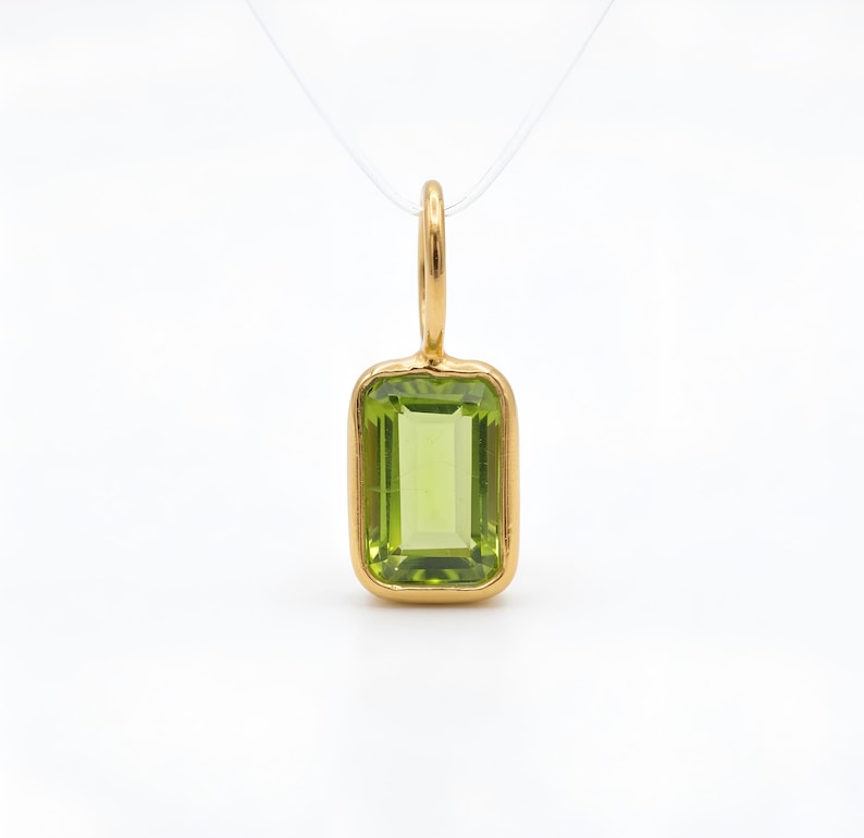 May include: A rectangular peridot pendant with a gold-coloured bezel and bail. The light green peridot stone is set in a gold frame. The pendant hangs from a thin, clear cord, and is set against a white background.