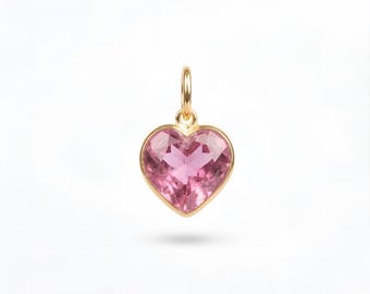 May include: A heart-shaped pendant featuring a faceted pink gemstone set in a polished gold-tone metal frame. The pendant includes a small circular gold-tone jump ring at the top for attachment to a chain or cord, showcasing a clear, bright pink hue.