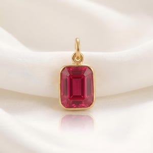 May include: A gold-colored pendant with a rectangular, ruby-red gemstone. The pendant has a simple, elegant design, with a polished finish. The gemstone is set in a gold bezel and has a faceted surface.