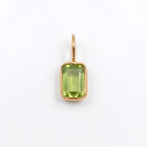 May include: A rectangular, emerald-cut light green gemstone pendant set in a polished gold-toned metal bezel. The pendant features a small, circular gold-toned metal bail at the top for attachment to a chain or cord.