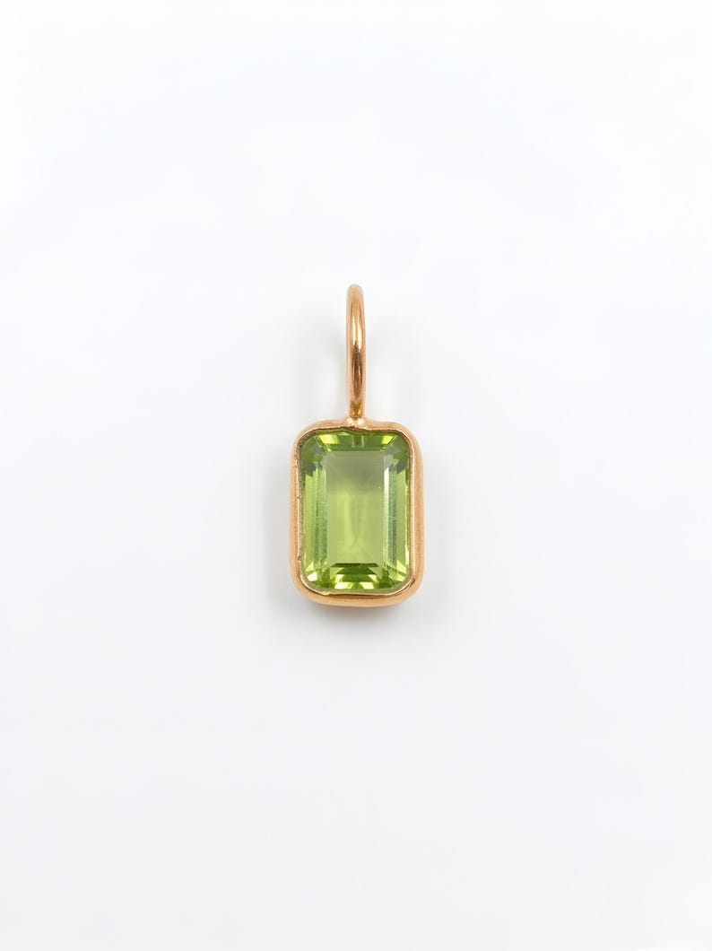 May include: A rectangular, emerald-cut light green gemstone pendant set in a polished gold-toned metal bezel. The pendant features a small, circular gold-toned metal bail at the top for attachment to a chain or cord.