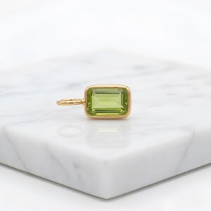 May include: A gold pendant with a rectangular, light green gemstone. The gemstone is set in a gold bezel and has a small loop for a chain. The pendant is displayed on a white surface.