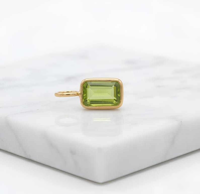 May include: A gold pendant with a rectangular, light green gemstone. The gemstone is set in a gold bezel and has a small loop for a chain. The pendant is displayed on a white surface.