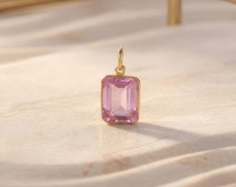 May include: A gold pendant featuring a rectangular, pale pink gemstone. The pendant has a small loop for a chain. The gemstone is faceted, reflecting light. The background is a neutral, textured surface.