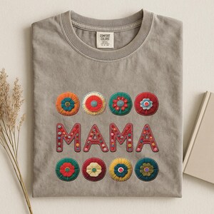 May include: A taupe Comfort Colors t-shirt with the word "MAMA" in red, embroidered letters. Above and below the word are colorful, circular floral designs in orange, red, green, and yellow.