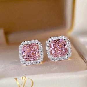 May include: A pair of square-cut pink gemstone stud earrings with a halo of smaller, clear gemstones. The earrings are set in a silver-colored metal and are displayed on a neutral background.