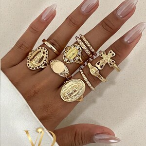 May include: A selection of gold rings on a hand. The rings showcase diverse designs, including religious motifs, decorative patterns, and a plain signet ring. The rings vary in size and style, creating a layered appearance. The image also shows the text "VALGUS DESIGNS".