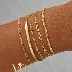 Puede incluir: Una variedad de pulseras doradas, cada una con un diseño único, adornan una muñeca. Las pulseras incluyen una cadena de cuerda trenzada, una cadena plana de espiga, una cadena con pequeñas cuentas y una cadena con eslabones ovalados. La imagen muestra el logotipo "VALGUS DESIGNS".