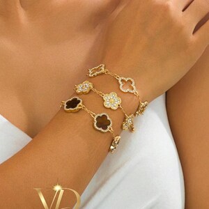 Pode incluir: Pulseira dourada com berloques em forma de trevo e flor. Alguns berloques são adornados com cristais transparentes, enquanto outros têm um centro marrom escuro. A pulseira é exibida no braço de uma pessoa.