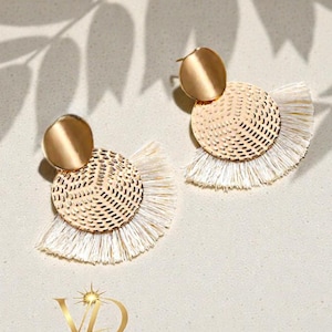 Może przedstawiać: Para złotych kolczyków o okrągłym wzorze i białych frędzlach. Każdy kolczyk ma mały, okrągły kołek i większe, teksturowane koło z tkanym wzorem. Neutralne tło z cieniami liści.