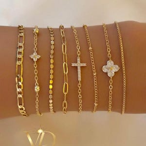 May include: A collection of gold-toned bracelets in various designs, including chain links, beaded, and charm styles. Some bracelets feature sparkling embellishments, such as a cross and a four-leaf clover. The bracelets are displayed on a light background.