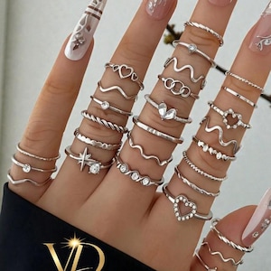Puede incluir: Un conjunto de anillos plateados con diversos diseños, incluyendo corazones, ondas y bandas trenzadas. Algunos anillos presentan pequeñas piedras preciosas. Los anillos se muestran en varios dedos, mostrando la variedad de estilos. El logotipo de la marca "VALGUSDESIGNS" es visible.